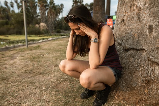 A woman kneels by a tree holding her head, expressing stress or anxiety in an outdoor park.